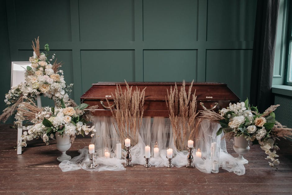 A somber funeral arrangement displayed on a wooden surface with a polished, dark brown casket centered behind it. Flanking the casket are two large white ceramic vases filled with cream and white rose