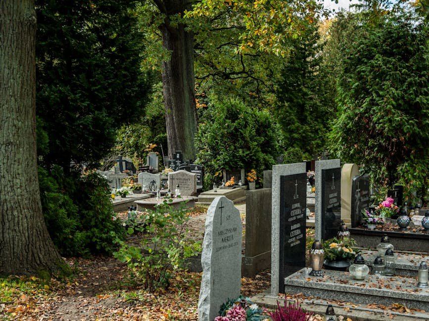 A serene cemetery scene featuring various graves adorned with fresh floral arrangements. Prominent in the foreground is a light gray headstone with inscribed text, surrounded by small flowering plants