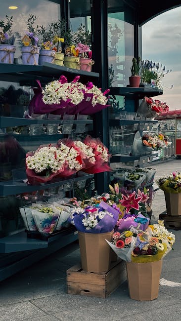 An outdoor display of various fresh floral arrangements and bouquets arranged on metal shelves and wooden crates, featuring a mix of colorful flowers including white, pink, purple, and yellow blooms.