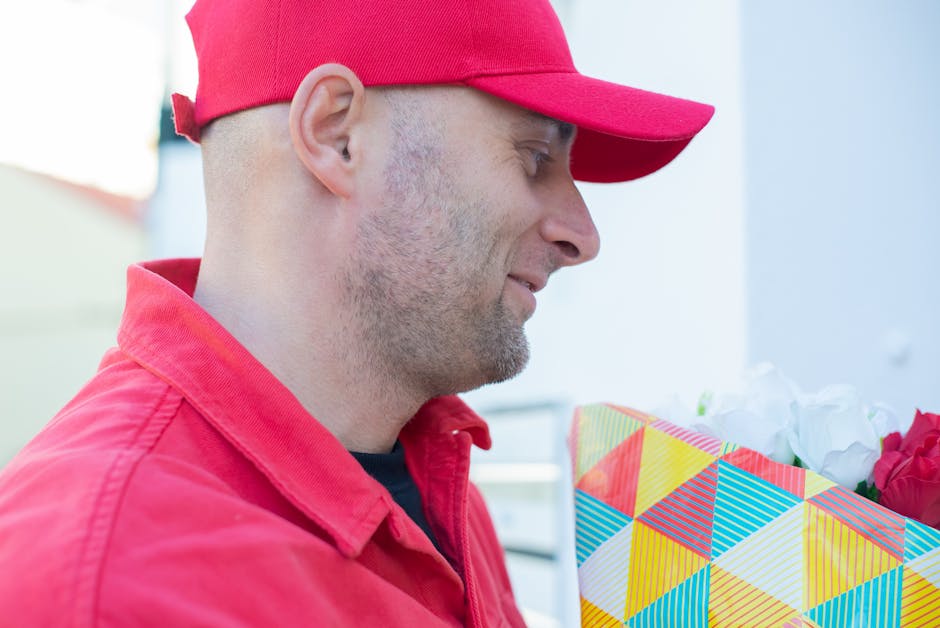 A man dressed in a red cap and red shirt holding a floral gift wrapped in colorful geometric-patterned paper, with white tissue paper and red flowers visible inside the wrapping. He is outdoors, stand