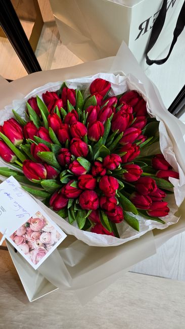 A large bouquet of vibrant red tulips with green stems and leaves, arranged in a dense and lush cluster. The flowers are wrapped in white tissue paper and placed inside a white gift box, which is laid