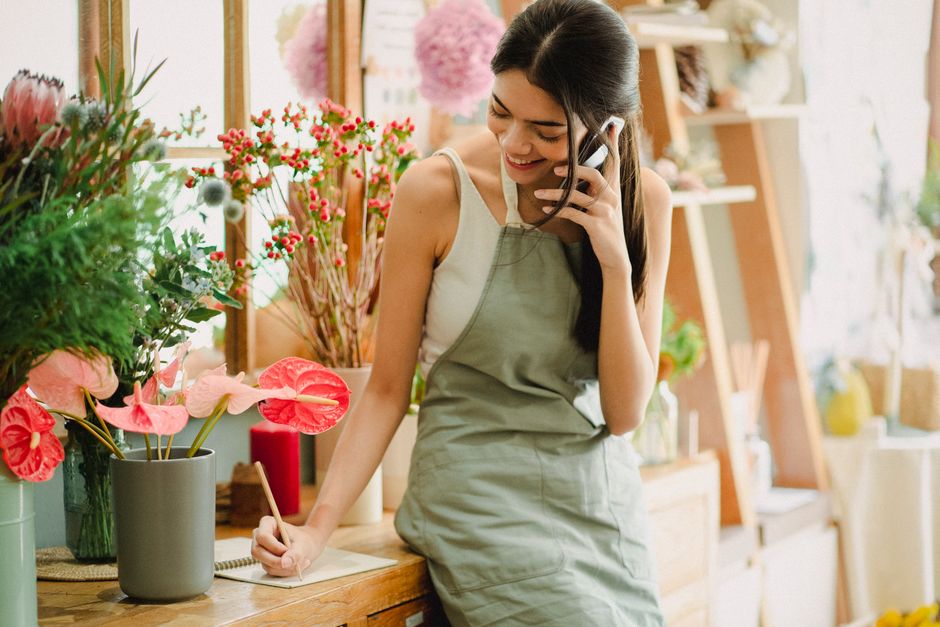A young woman with dark hair, smiling, wearing a light-colored sleeveless top and a beige apron, stands inside a flower shop. She is holding a landline phone with her left hand close to her ear and wr