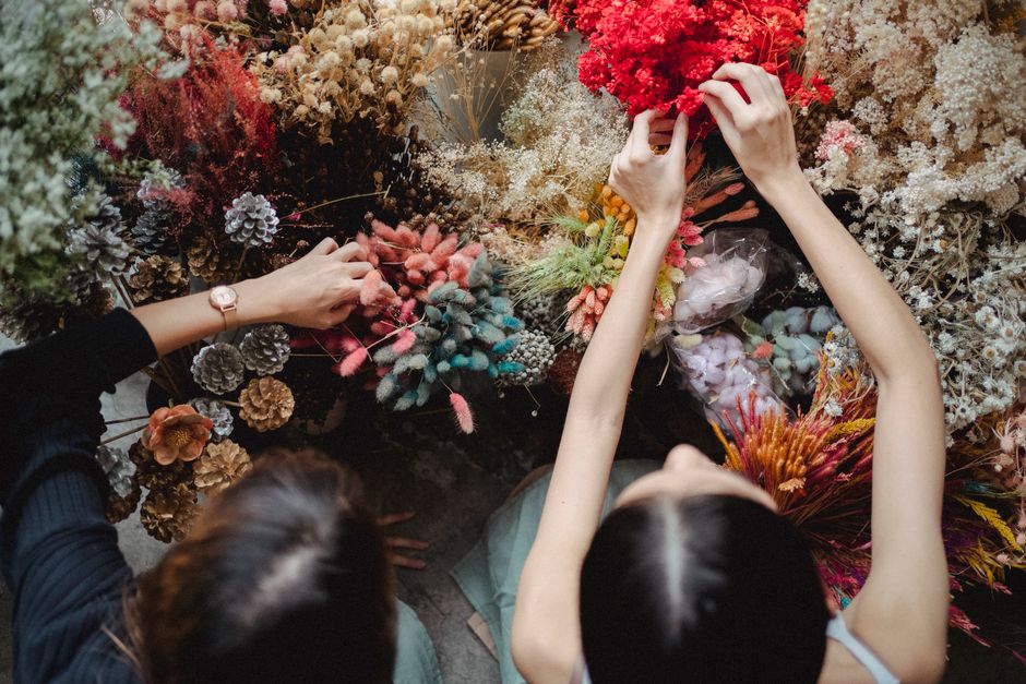 Two floral designers are arranging a vibrant bouquet of mixed flowers on a table surrounded by an array of dried and fresh blossoms, including pink, red, white, and purple blooms, as well as pinecones
