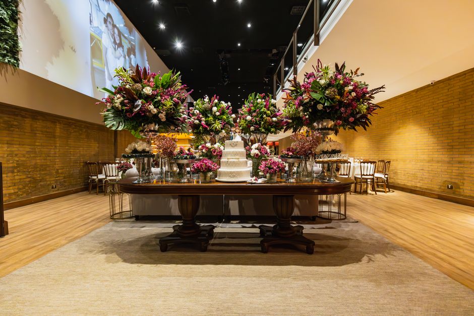 A large, elegant floral arrangement featuring pink, purple, and white flowers with green foliage is displayed on a polished wooden table in a spacious hall with warm brick walls and wooden flooring. T