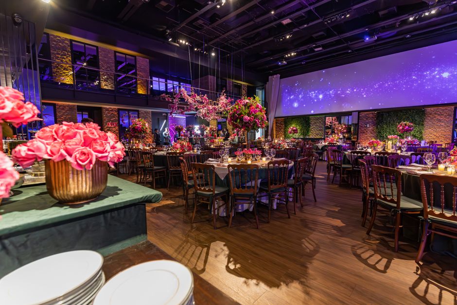 An elegant event hall decorated with an array of vibrant floral arrangements, including large bouquets of pink roses and peonies in copper and glass vases placed on tables covered with white tableclot